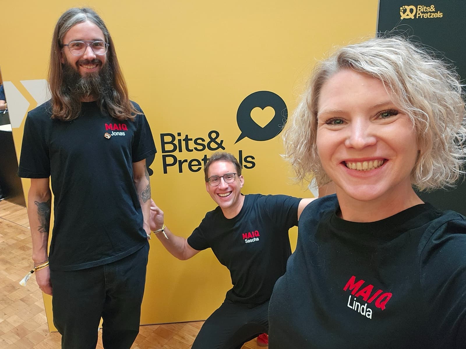 Jonas, Sascha and Linda in black MAIQ shirts in front of the yellow Bits & Pretzels wall