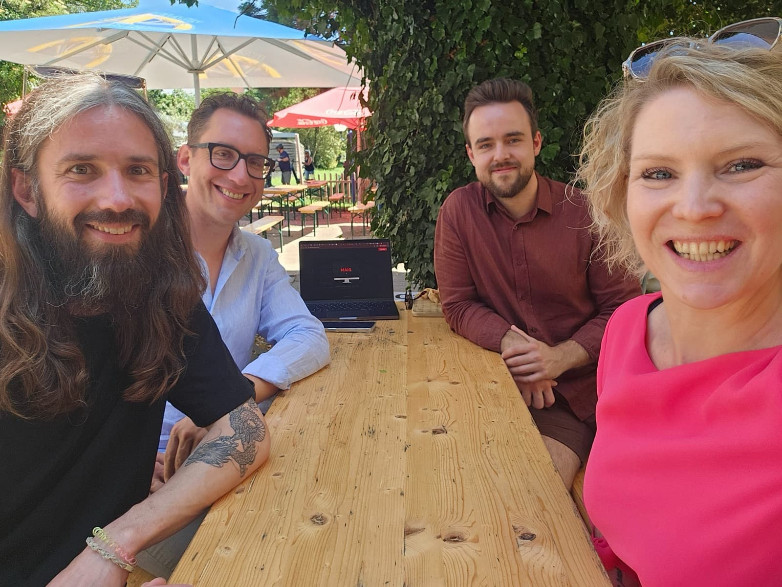 Jonas, Sascha, Eike and Linda at a wooden table in a beer garden, a laptop with the MAIQ logo in front of them
